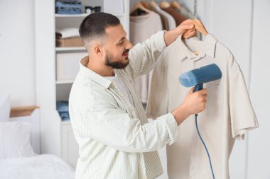Genç adam yatak odasında buharlı gömlek giyiyor.