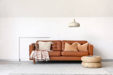 Beyaz duvarın yanında minderleri ve pufları olan kahverengi deri kanepe.