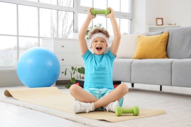 Evde yoga minderinde oturan halterli mutlu küçük çocuk.