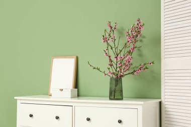 Üzerinde fotoğraf çerçevesi, giyinme ekranı ve yeşil duvarın yakınındaki pembe çiçek açan vazolardan oluşan bir elbise dolabı.