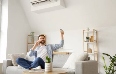 Genç adam cep telefonuyla konuşuyor ve oturma odasında klimayı açıyor.