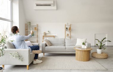 Genç adam oturma odasında, arka manzaralı kanepede klimayı açıyor.