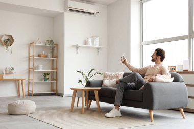 Genç adam evdeki kanepeden klimayı açıyor.