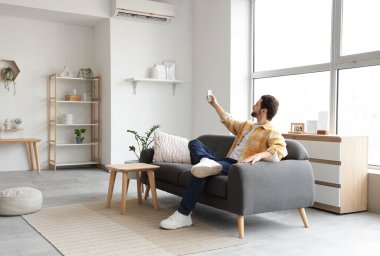 Genç adam evdeki kanepeden klimayı açıyor.