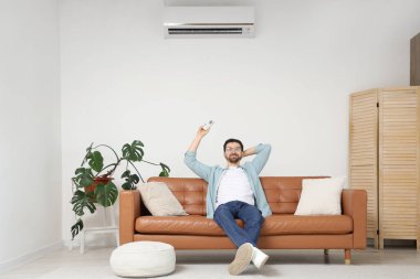 Yakışıklı adam oturma odasındaki kanepeden klimayı açıyor.