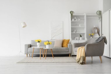 Oturma odasının içinde gri kanepe, koltuk ve vazolarda nergisler var.