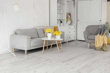 Oturma odasının içinde gri kanepe, koltuk ve vazolarda nergisler var.