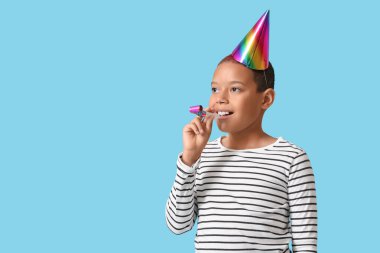 Parti şapkalı şirin Afro-Amerikalı çocuk mavi arka planda doğum gününü ıslıkla kutluyor.