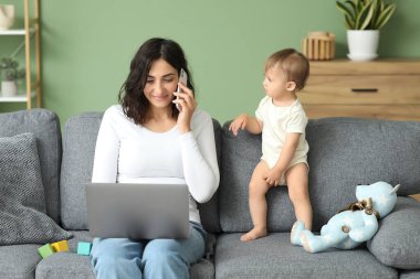 Evde cep telefonuyla konuşurken dizüstü bilgisayarla çalışan tatlı bir bebek ve genç anne.