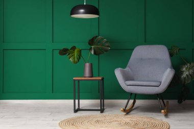 Yeşil duvarın yanındaki kahve masasında canavar yaprakları olan gri koltuk ve vazo.