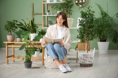 Evdeki çiçeklerin yanında pouf hakkında kitap okuyan genç bir kadın.