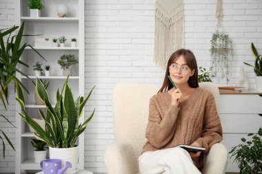 Evdeki çiçeklerin yanındaki koltukta oturan defterli genç kadın.