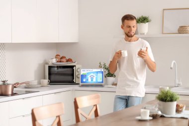 Elinde kahve fincanı olan genç adam mutfaktaki cep telefonunda akıllı ev uygulaması kullanıyor.