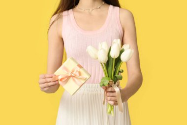 Sarı arka planda Anneler Günü için hediye kutusu ve güzel lale çiçekleri tutan genç bir kadın.