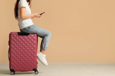 Bej duvarın yanında bavulu ve cep telefonu olan genç bir kadın.