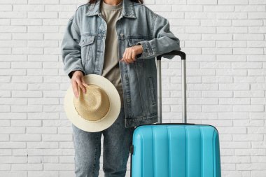 Beyaz tuğla arka planda bavulu ve şapkası olan genç bir kadın.