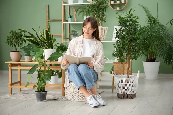 Evdeki çiçeklerin yanında pouf hakkında kitap okuyan genç bir kadın.