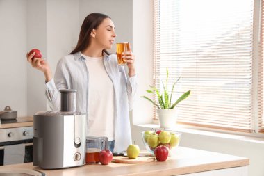 Modern meyve sıkacağı olan genç bir kadın mutfakta elma suyu içiyor.
