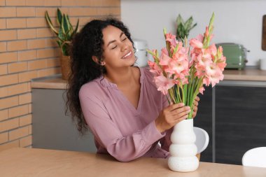 Güzel, mutlu, Afro-Amerikan bir kadın. Elinde pembe gladyatör çiçeği buketi var. Mutfakta oturuyor.