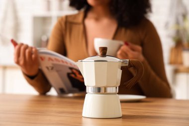Elinde bir fincan taze espresso ile mutfakta moda dergisi okuyan genç bir kadın gayzer kahve makinesine odaklanıyor.