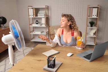 Elektrikli yelpazesi ve kokteyli olan güzel, mutlu genç bir kadın iş yerinde oturuyor.