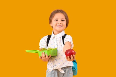 Sırt çantası, beslenme çantası ve sarı arka planda elma olan mutlu küçük kız.