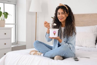 Güzel, Afro-Amerikalı bir kadın, elinde bir sepet dondurmayla evde, yatağında oturuyor.