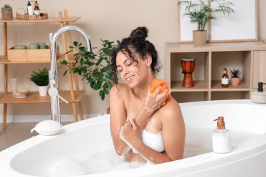 Genç mutlu Afro-Amerikalı kadın evde sünger, duş jeli ve sabun köpüğüyle banyo yapıyor.