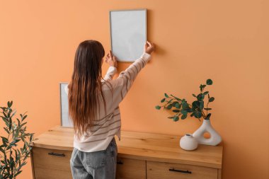 Evdeki turuncu duvara boş bir çerçeve asan genç bir kadın.