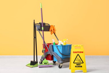 Temizlik malzemeleri ve sarı duvarın yanında uyarı levhası olan kapıcı vagonu.