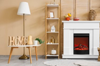 Bej duvarın yanındaki modern şöminede raf, masa ve sonbahar süslemeleri olan şık bir oda.