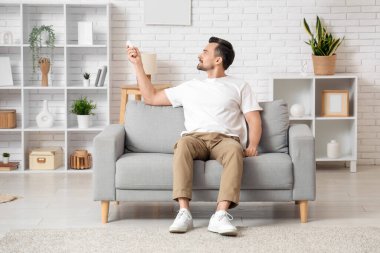 Beyaz duvarın yanındaki kanepede oturan klimalı uzaktan kumandalı yakışıklı genç adam.