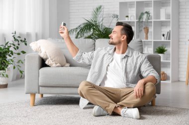 Klima kumandalı yakışıklı genç adam oturma odasında yerde oturuyor.