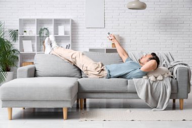 Klimalı uzaktan kumandalı genç adam beyaz tuğla duvarın yanındaki kanepede yatıyor.