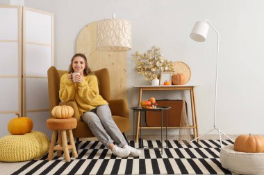 Güzel, genç ve mutlu bir kadın çay fincanı ile sonbahar için dekore edilmiş olarak evdeki koltukta oturuyor.