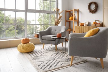 Sonbahar için dekore edilmiş modern oturma odasının içinde rahat koltuklar, sehpa ve pencere kenarında balkabakları var.