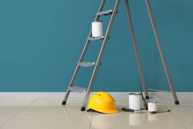 Mavi duvarın yanında boya, fırça ve baret bulunan merdiven.