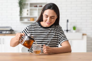 Güzel, genç, mutlu bir kadın mutfaktaki şeffaf çaydanlıktan bardağa sıcak çay dolduruyor.