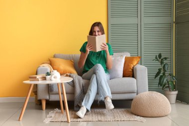 Sarı duvarın yanındaki kanepede kitap okuyan güzel bir kadın.