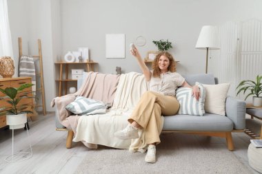 Oturma odasındaki kanepede oturan klimalı uzaktan kumandalı güzel, gülümseyen genç bir kadın.