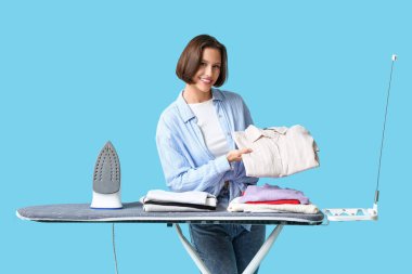 Mavi arka planda ütü masasında demir ve temiz giysiler yığını olan genç bir kadın.