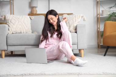 Pembe pijamalı kadın evde dizüstü bilgisayarla halıda çalışıyor.
