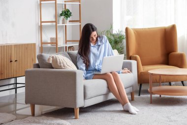 Evdeki kanepede dizüstü bilgisayarla çalışan mavi pijamalı genç bir kadın.