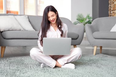 Pembe pijamalı kadın evde dizüstü bilgisayarla halıda çalışıyor.