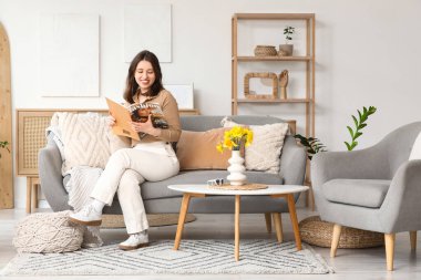 Elinde vazo dolusu nergis çiçeği olan mutlu genç bir kadın oturma odasındaki kanepede çiçek okuyor.