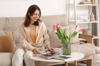 Lale çiçekleri ve dergilerle dolu vazosuyla oturma odasındaki kanepede oturan mutlu genç kadın.