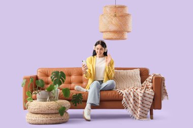 Kulaklıklı genç bir kadın kahverengi koltukta cep telefonu kullanıyor.