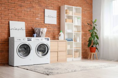 Çamaşır odasının içinde, modern çamaşır makinesi ve kurutma makinesi olan tuğla duvarın yanında.