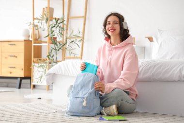 Yatak odasında sırt çantasından kitap alan kulaklıklı Hintli bir kız öğrenci.