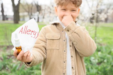 Hasta küçük çocuk elinde burun damlası ve açık havada ALLERGY yazılı doku tutuyor.